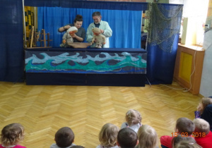 Scenografia teatralna - morze, dwóch aktorów z lalkami - rybakiem i żoną rybaka oraz naczyniem. Dzieci oglądające przedstawienie.